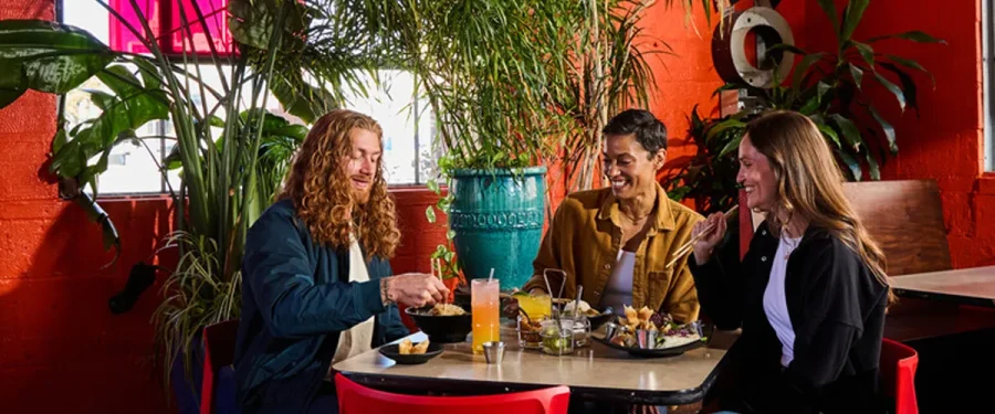 Three customers sit at a table and eat from an array of delicious food with various plants in the background.