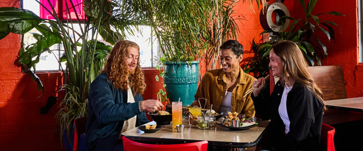 Three customers sit at a table and eat from an array of delicious food with various plants in the background.