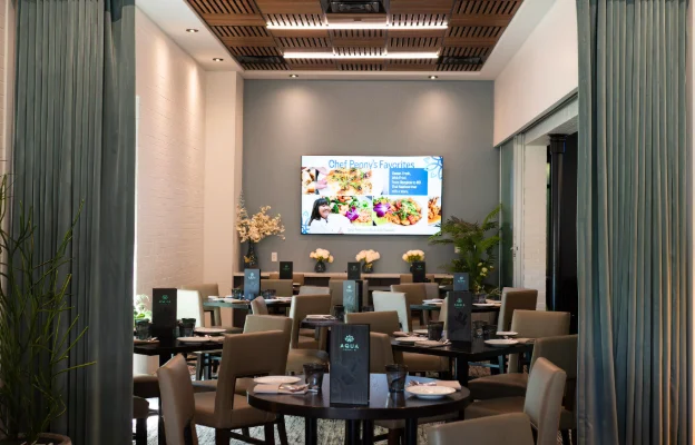 Large room at Aqua Penny’s with multiple table sets and chairs along with a large television against the back wall and two colored drapes.