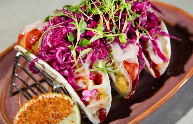 Three tacos on a taco stand filled with meat, cheese, sauce, and covered by pickled onions and veggies.