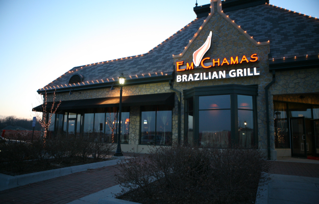 The outside of Em Chamas Brazilian Grill in Kansas City as the sun begins to set and a neon sign is lit up on the front of the restaurant.