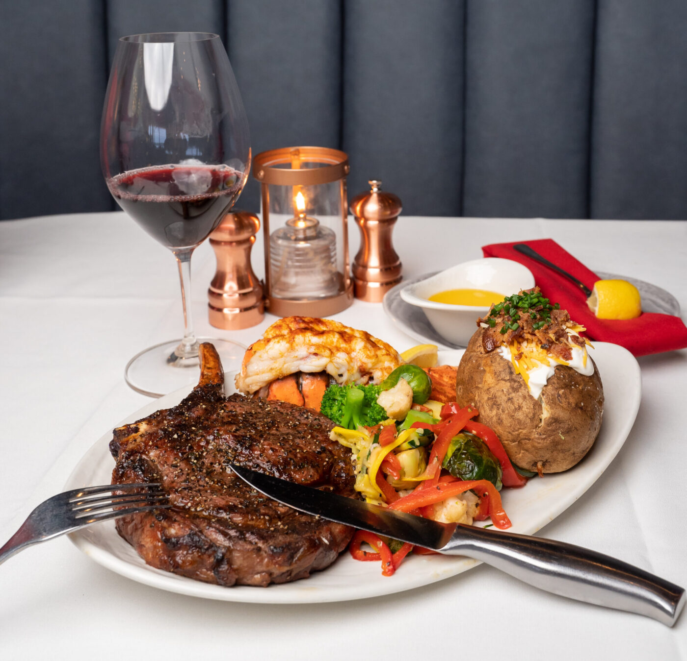 A gourmet surf-and-turf dinner with a grilled bone-in ribeye steak, baked and loaded potato, lobster, and veggies coupled with a wine glass.
