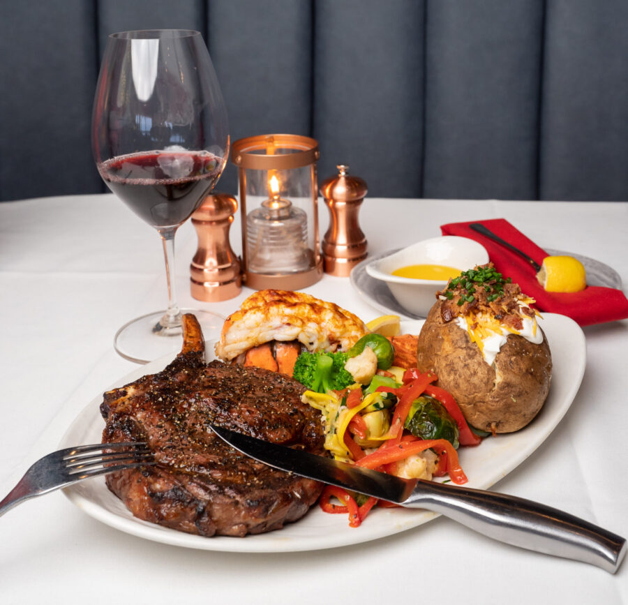 A gourmet surf-and-turf dinner with a grilled bone-in ribeye steak, baked and loaded potato, lobster, and veggies coupled with a wine glass.