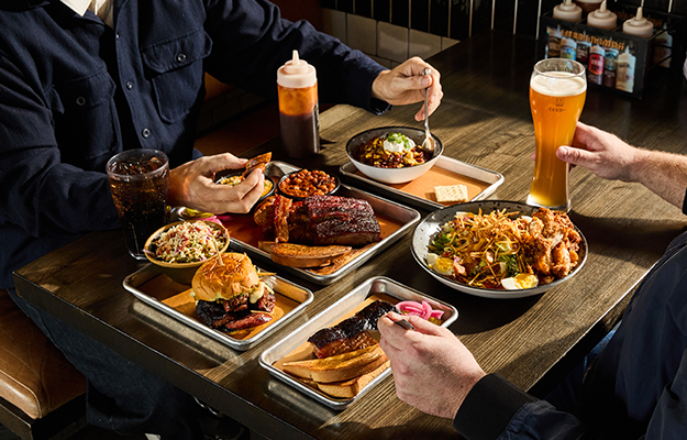 Two customers enjoy an array of Barbecue foods and beer including ribs, pulled pork sandwich, coleslaw, and more.