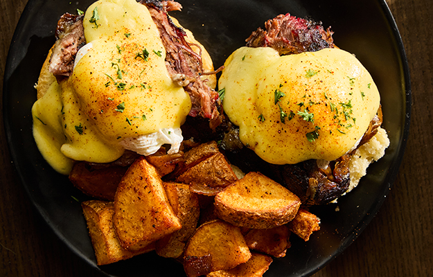 BBQ pulled pork eggs Benedict served on a black plate with potato wedges.