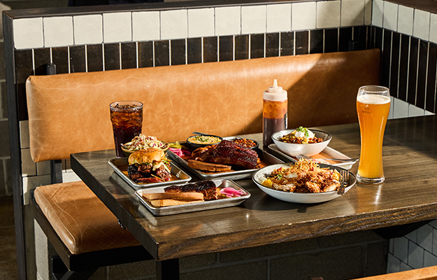 A table full of Barbecue foods and beer including ribs, pulled pork sandwich, coleslaw, and more.