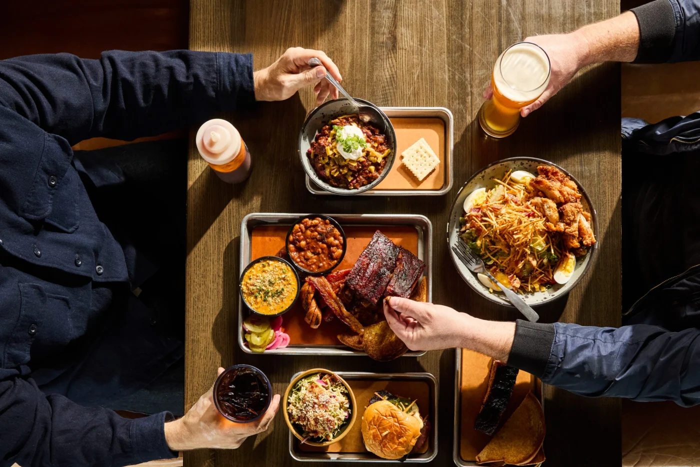 Two customers enjoy a spread of barbecue foods including beans, mac-and-cheese, pulled pork sandwich, ribs, and more.