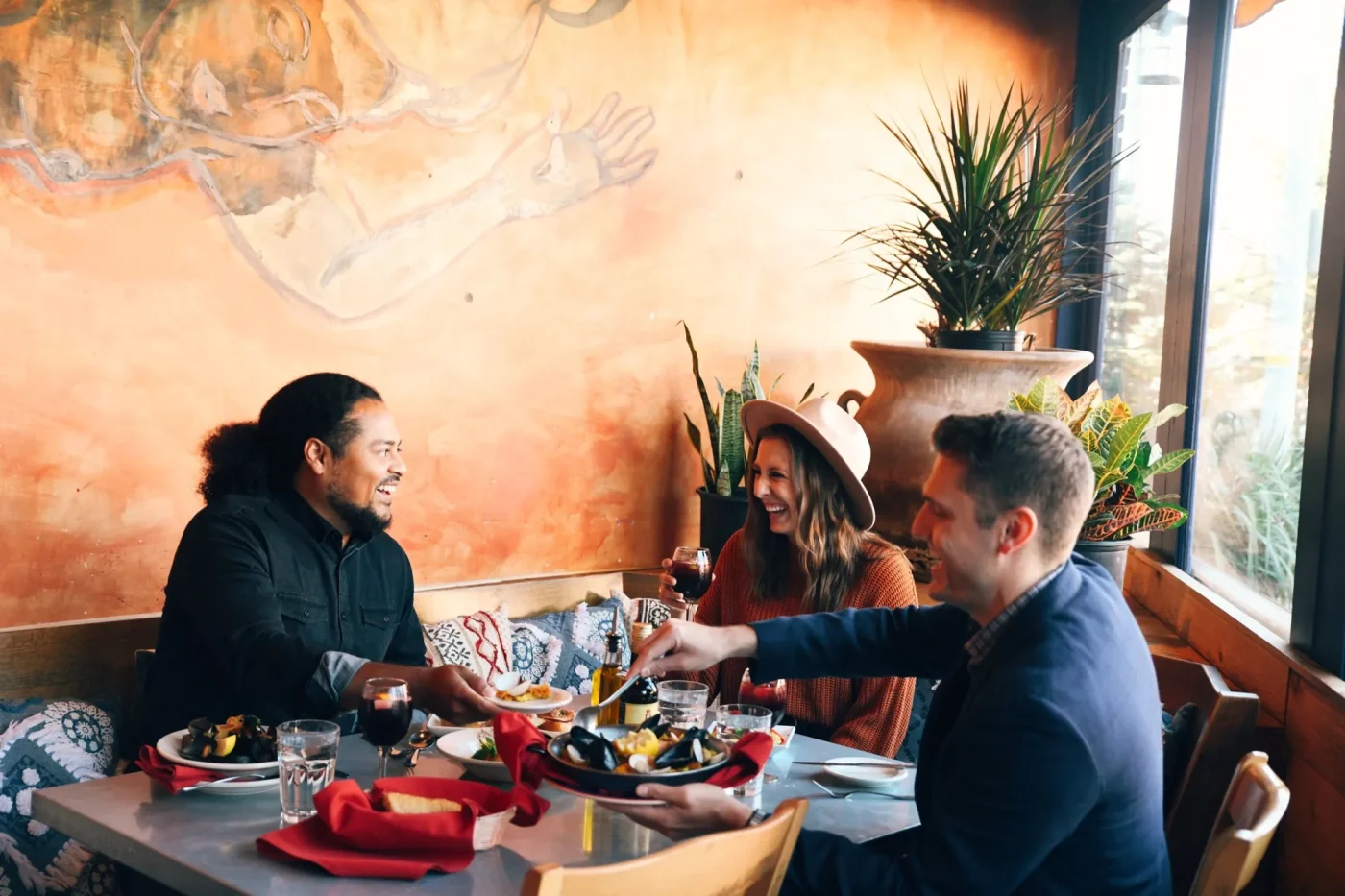 Three friends enjoy a spread of food at a restaurant in a corner booth with plants in the background.