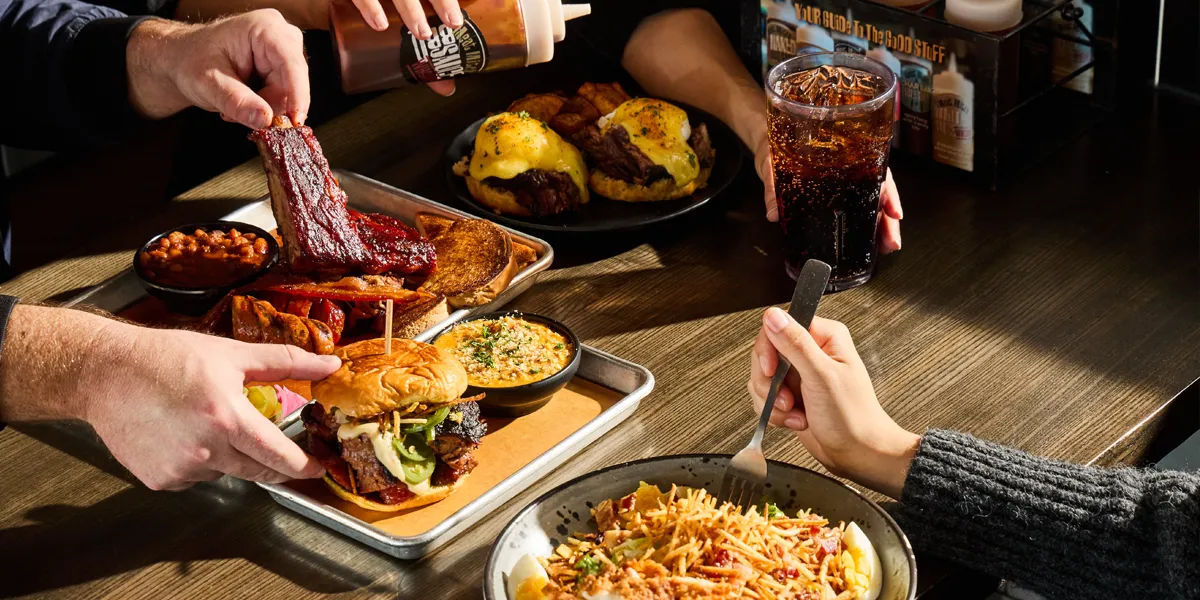 Three customers enjoying a spread of delicious BBQ food including burgers, ribs, toasted bread, and more.