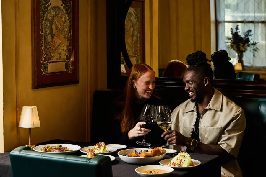 Beautiful couple at a restaurant tap their wine glasses together with a spread of delicious food on the table in front of them.