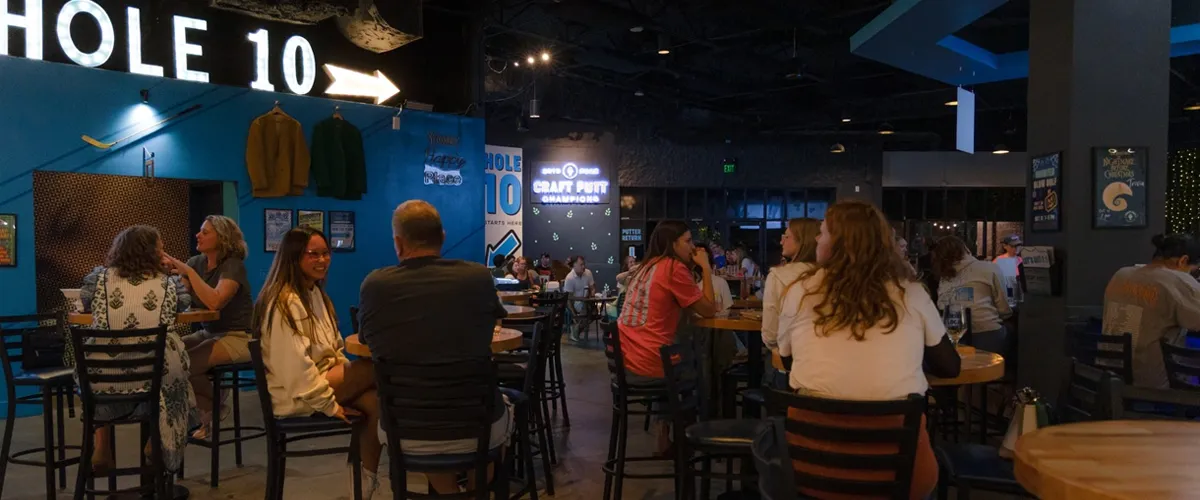 The interior of craft putt where various customers sit at wooden tables and talk while eating food and having fun.
