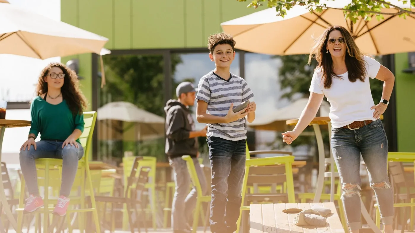 A family plays corn hole outside at a scenic venue with tables and umbrellas during a sunny day.