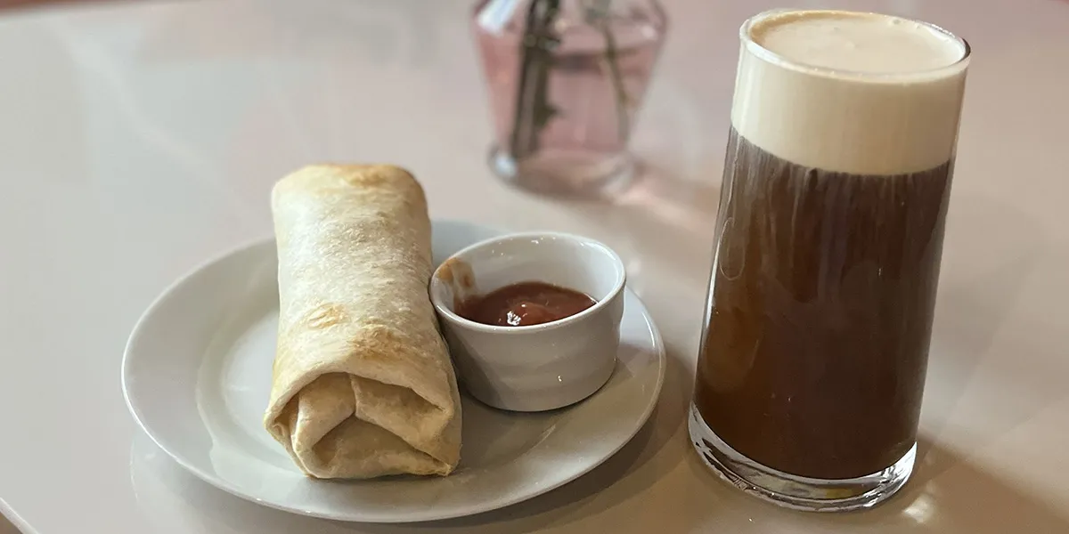 Two Sugars breakfast burrito on a small white plate served with sauce next to a caffeinated beverage.